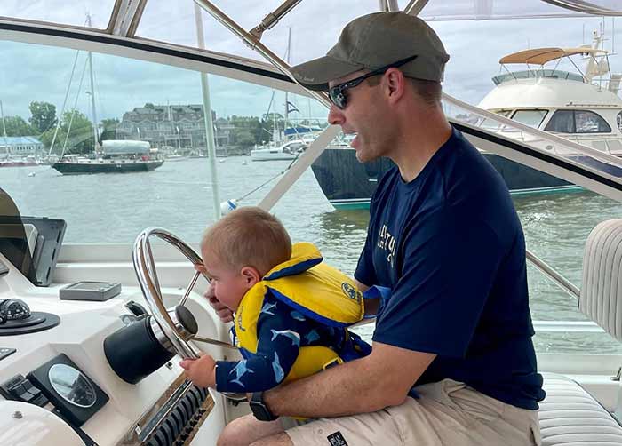 Man in a blue shirt, ball cap and sunglasses sitting at the hlem of a powerboat with baby in his lap
