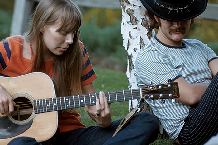 Young Joni Mitchell playing acoustic guitar next to David Crosby leaning against a tree