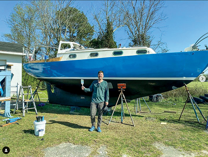 Photo of a young adult male holding a paint brush in front of a blue and white boat