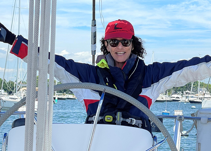 Middle-aged Caucasian female wearing sunglasses, a red baseball hat and a navy blue jacket at the wheel of a vessel on a sunny day