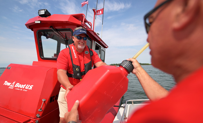 Middle-aged man wearing a red shirt, red life jacket and aboard a Tow BoatU.S. handing a gas tank to a man on another boat.
