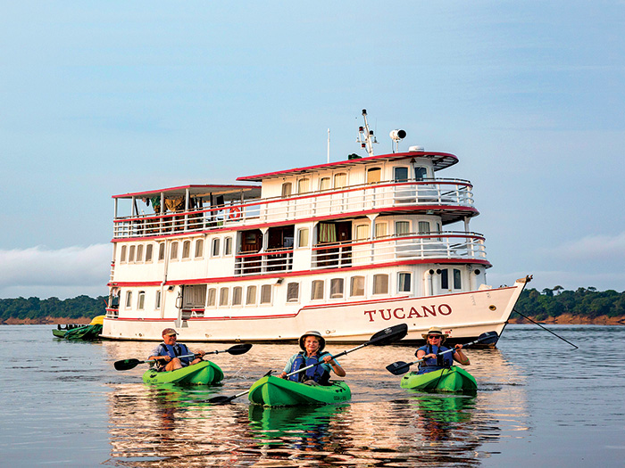 Three adults in green kayaks paddling away from a large white boat.