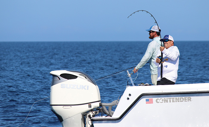 Two adult males wearing white and blue hats fishing off the back of a white boat in open waters.