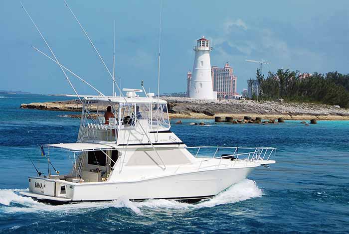 a large white sportfishing boat is underway near land with a white lighthouse and tall high-rise buildings in the distance.