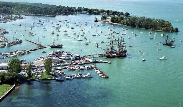 areal photograph of a small bay area with numerous boats in the water and several large boats with masts. 