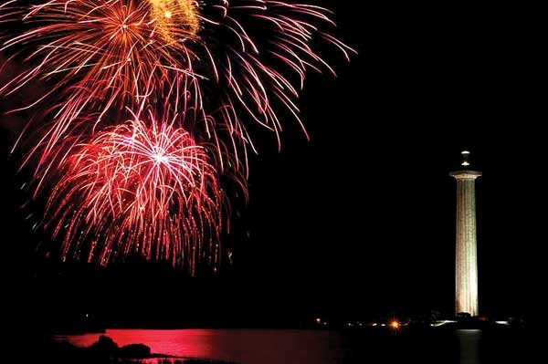 Several red fire works exploding in the air under a night sky across from a white lighthouse