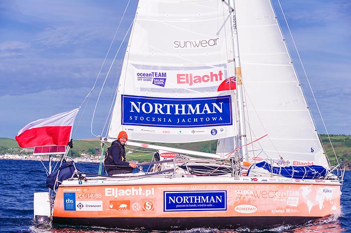 A man smiles from the helm of his sailboat. The sailboat and sails are covered in various logos.