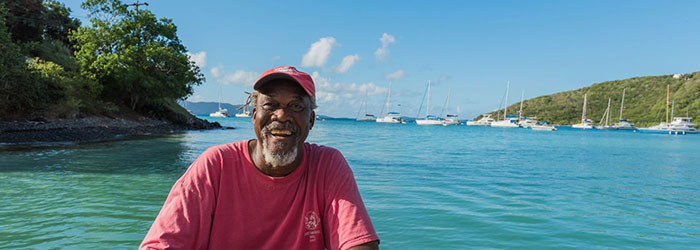 A man smiles at the camera wearing a red hat with turquoise waters behind him and boats moored in the distance