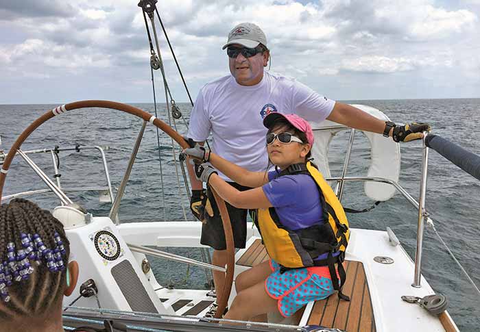 A man with his daughter at the helm of a sailboat. They are both wearing a hat, sunglasses and sailing gloves.