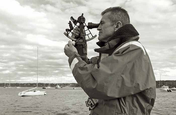 Black and white photo of man holding a sextant up to his eye to navigate at sea