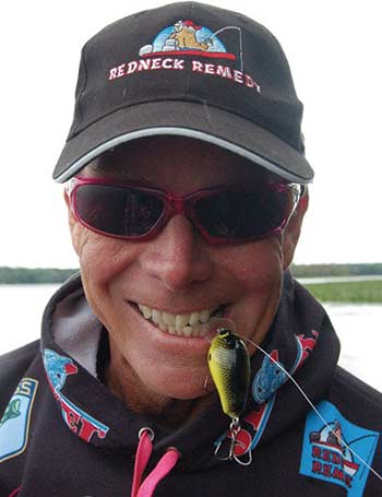 A man wearing a baseball hat and red sunglasses holds a green fishing lure in his teeth