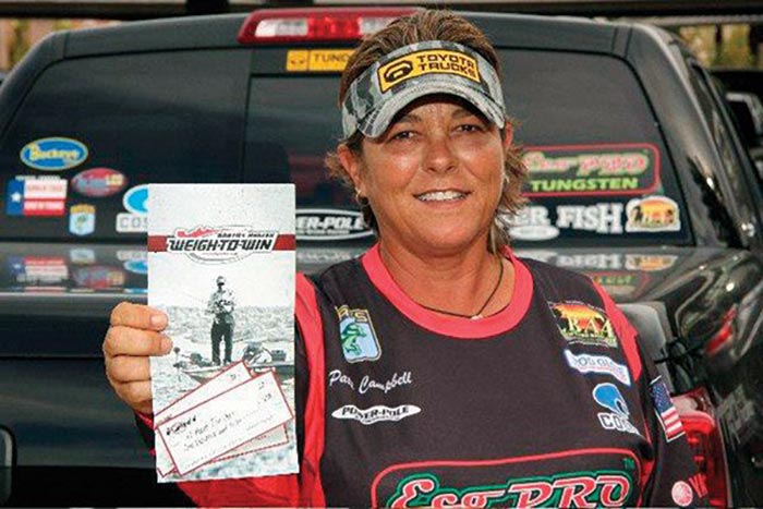 Woman wearing camoflauge visor smiles while holding up a fishing brochure and standing in front of a truck full of fishing stickers