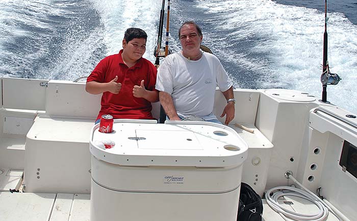 Man in a white shirt sitting next to a boy in a red shirt cruising on a sailboat with fishing rods behind them