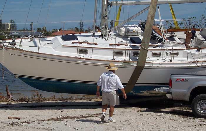 A man with  the words "BoatUS" on the back of his shirt walks toward a sailboat held in straps that is on land