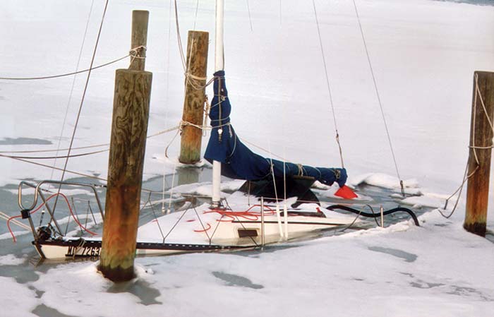 Sailboat tied to piles sinking into to ice and snow with only to top of the boat and the mast visible
