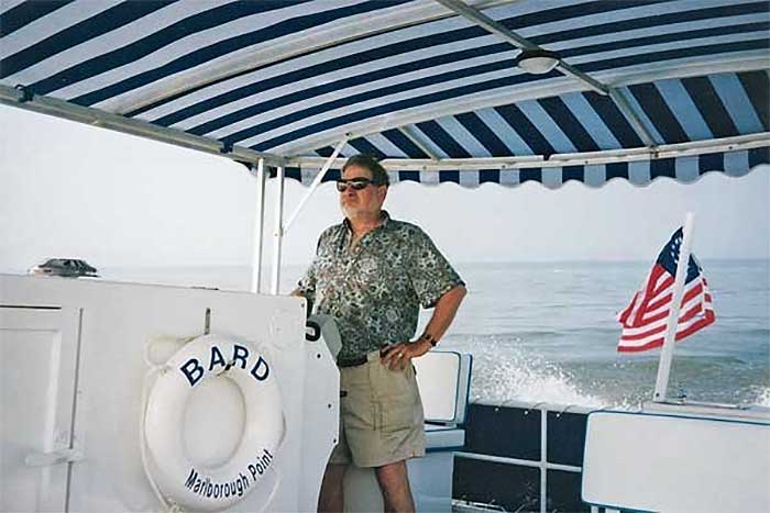 Man wearing a pair of tan shorts and printed shirt with sunglasess standing at the helm of a large pontoon boat