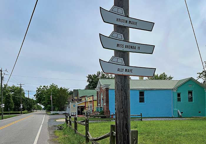 A wood pole with boat-shaped sign attached to it, with a wooden fence, road and houses in the background