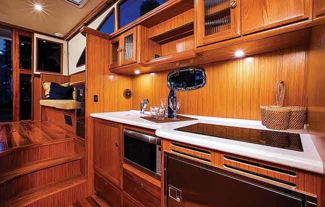 A multi level brown teak wooden interioir of a boat, showing a cook top and oven as well as many storage cabinets. Up the three stairs is a small sitting space with a pillow and glass windows.