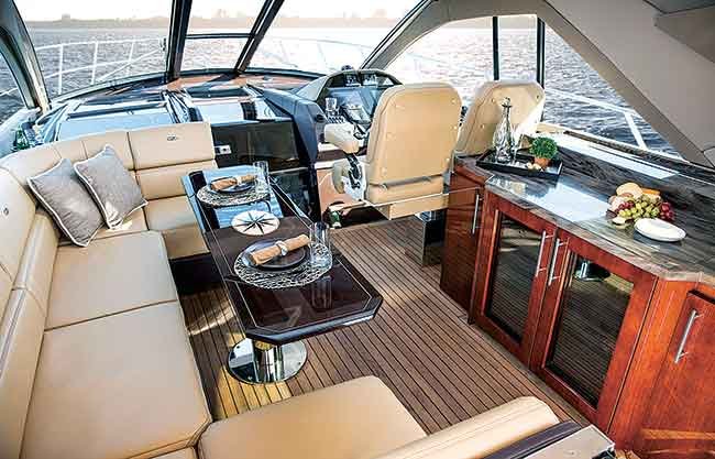  A beige L-shaped couch, helm station and wooden table make up the Interior of Regal 53 Sport Coupe powerboat salon. Dishes and cloth napkins decorate the space.