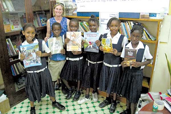 a white woman with blond hair poses with a group of 6 black children dressed alike in school uniforms holding up books