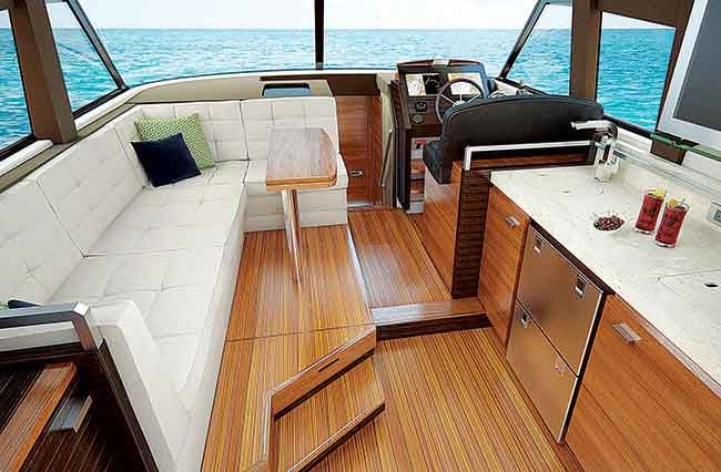 Salon seating section of boat, modern straight lines with a L-shaped white sitting area, and small helm station. Two glasses of ice tea sit atop a sparkling white countertop. Open blue water viewable out front windows.