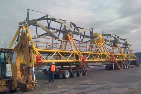 Large yellow metal structure on top of a large trailer. Man in bright orange construction vest and white hard hat looks up at rig. Yellow forklift in foreground.