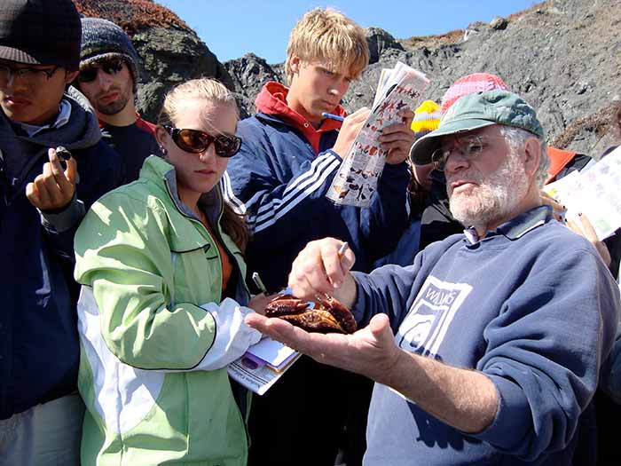 A group of students gather around a man holding a crab. One student holds up and writes on a colorful legend of sea creatures, including crabs.