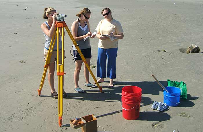 Three women on beach using survey equipment, surrounded by several buckets and equipment. 