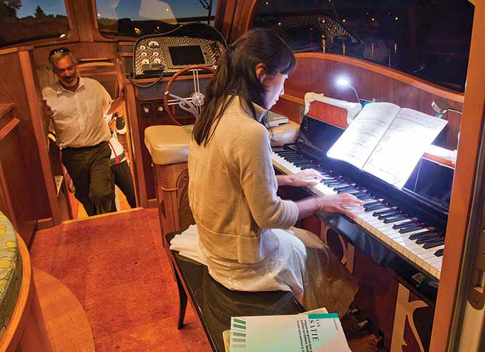 Lionheart's Concerto yacht interior with young girl playing the electric Yamaha player piano and a man standing in the cabin watching