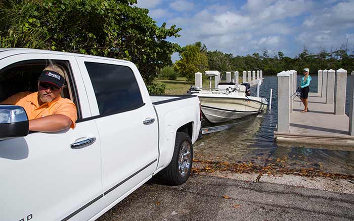 Man in white pickup truck looking in side mirror watching the boat on its trailer as the boat is halfway in the water at a boat ramp
