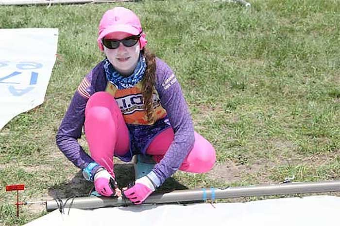 Young girl wearing a purple top, pinking leggings, sunglasses and a  pink ballcap ties sail to mast on ground