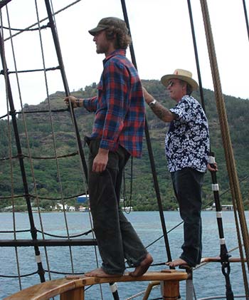 Two man wearing hats standing in sailboat rigging with water and coastline in the background