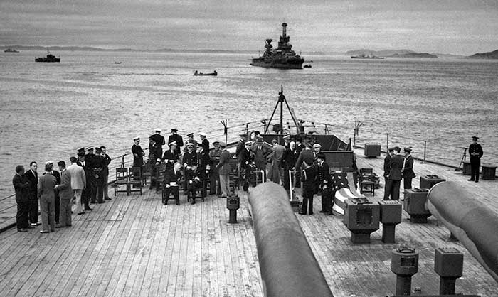 Big guns of the HMS Prince of Wales, overlooking a gathering of people including FDR and Churchill seated in front