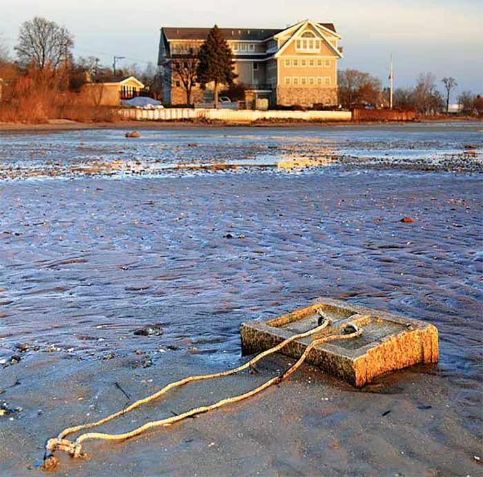 A mooring that once sat several feet underwater lies exposed in shallow water with large house in the background