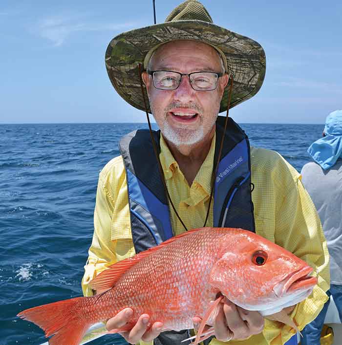 Older man in hat and wearing glasses holding a large red snapper at sea