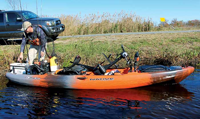 Man loading fishing gear in a kayak at the water edge with truck in the background