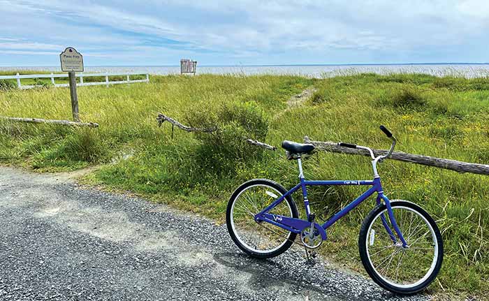 A blue bicycle sitting on the side of a gravel road with a sign that says "Photo Stop" and grass field with body of water in the background
