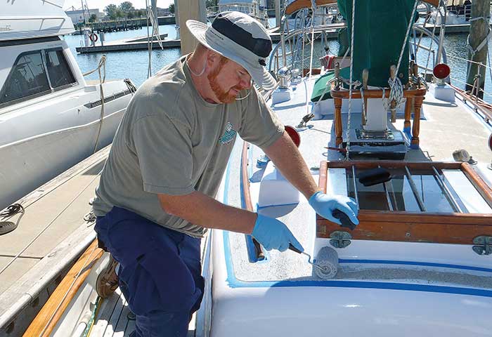 Man wearing blue work pants, gray shirt, sunhat, also wearing rubber gloves while standing and painting hull with a roller brush