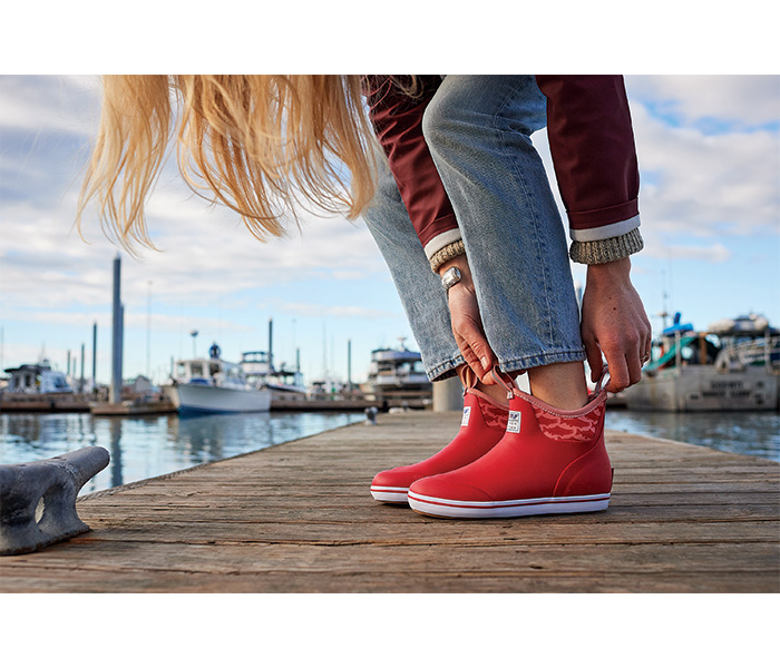 Adult female standing on a dock putting on red ankle boots.