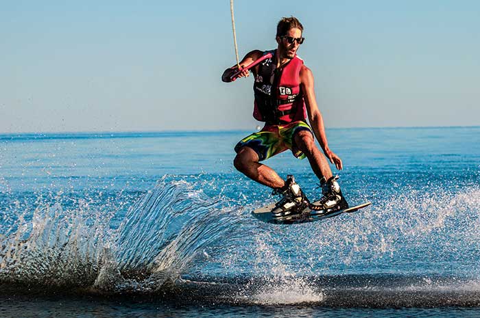 Phot Contest Action & Watersports Finalist: Image of man on a wakeboard wearing an orange lifejacket and sunglasses jumping over a wake on the water