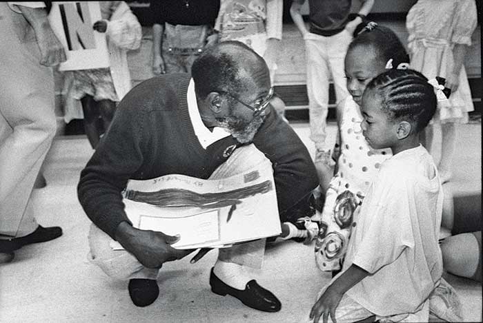 Black and white photo with an older man kneeling down holding open a children's book with two little girls kneeling beside him