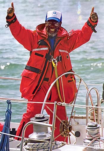 Older man standing on a sailboat with his hands in the air wearing orange fishing waders and and orange jacket