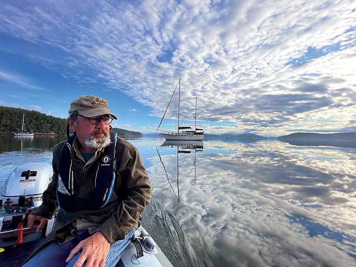 Photo Contest Boating Lifestyle Winner: Image of a man sitting in a dingy hand on the tiller crusing through the water with a sailboat in the background