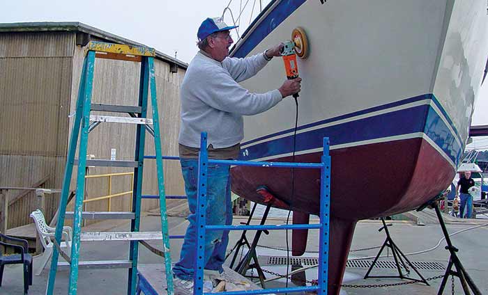 Man in jeans, sweatshirt and ball cap, standing by boat on a jackstand and using am electric buffer on hull