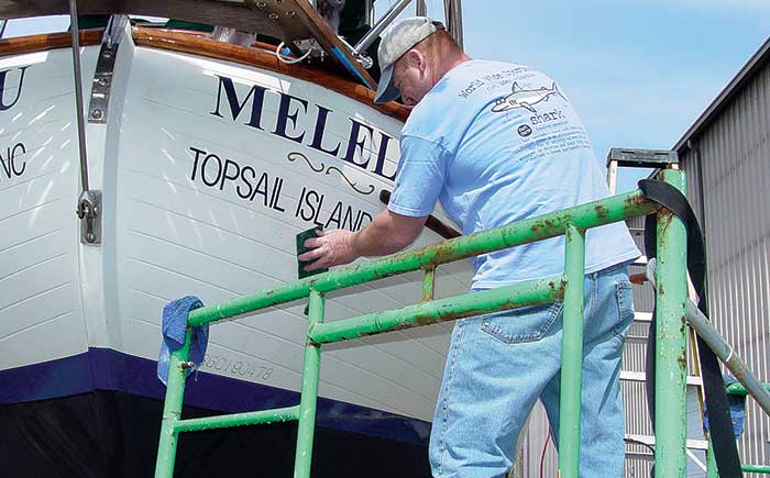Man wearing jeans, blue t-shirt and white ball cap standing on scaffolding and cleaning boat hull withscrubbing pad