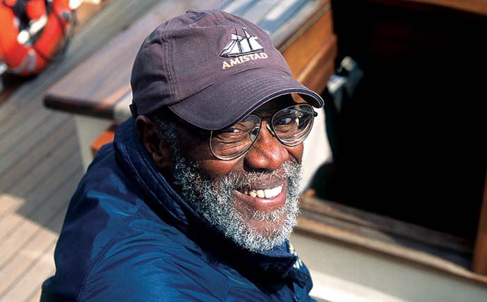 Portrait of an older man wearing a blue jacket, blue ball cap, beard and mustache and glasses smiling at the camera