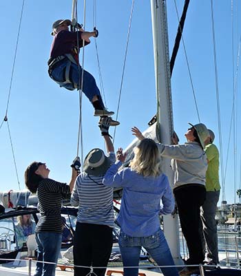 Woman climbing the mast of a sailboat while a group of women cheer her on at the base of the mast