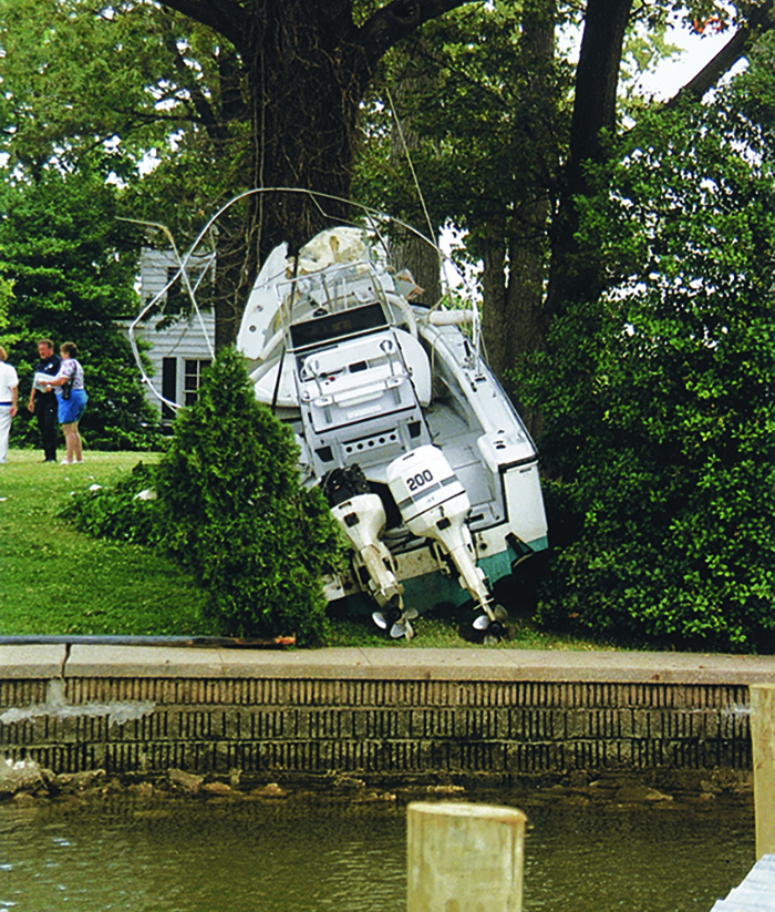 White boat that jumped a sea wall and crashed into a tree and bushes in front of a white house.