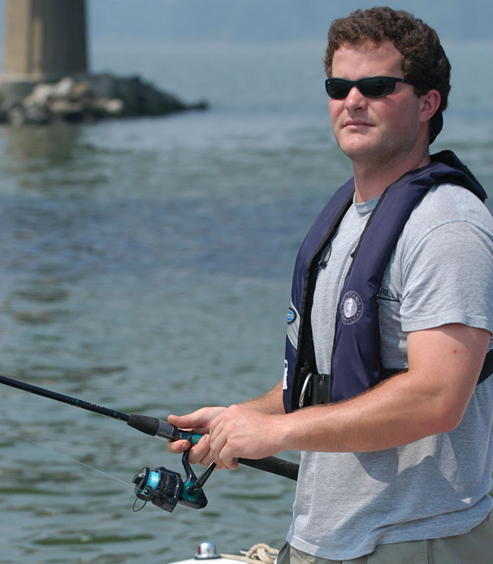 Young adult Caucasian male wearing sunglasses, gray shirt and a navy life jacket fishing off a boat.