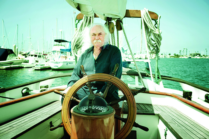 David Crosby in his later years wearing a black collared shit at the wheel of a sailboat on a sunny day